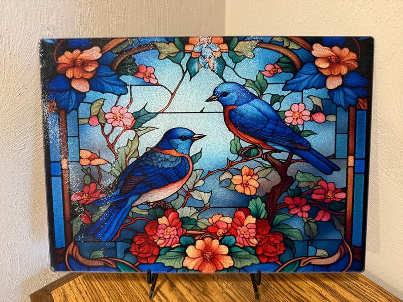 A square glass cutting board with a stained-glass style image of two bluebirds perched on flowering branches, set against a light blue background.