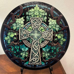 A round cutting board with a Celtic cross design in the center, surrounded by green leaves and vines, resembling stained glass, displayed upright on a black stand.