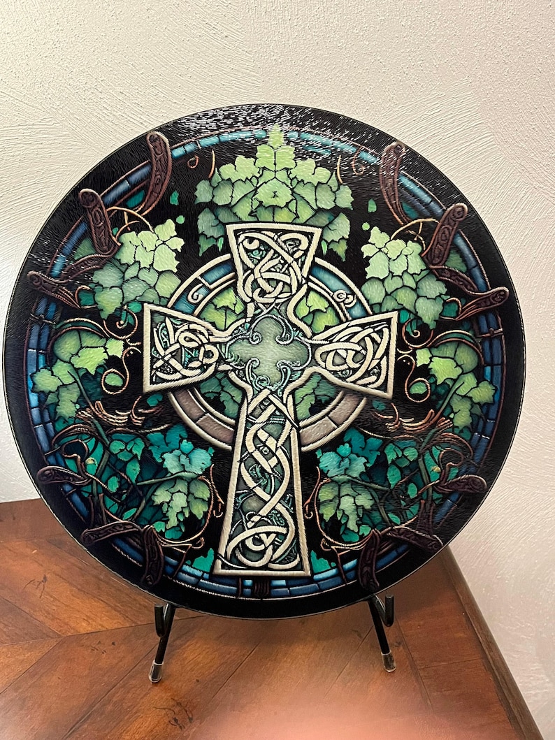 A round cutting board with a Celtic cross design in the center, surrounded by green leaves and vines, resembling stained glass, displayed upright on a black stand.