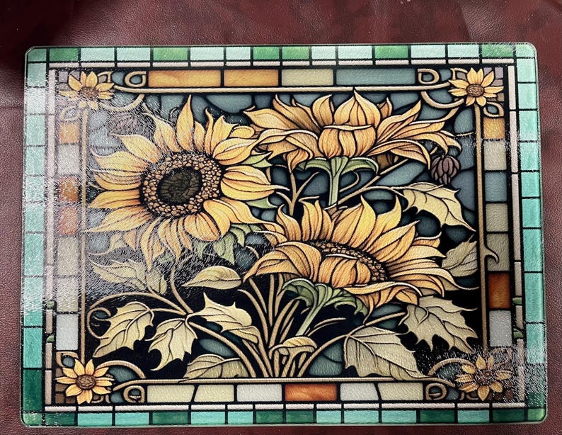 May include: A glass cutting board with a stained glass design featuring sunflowers in shades of yellow, brown, and green. The board is framed with a mosaic of blue, green, and orange tiles.