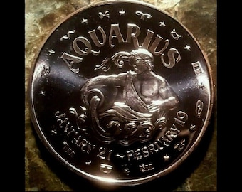 Peut inclure: Une pièce de monnaie du zodiaque Verseau de couleur argentée. La pièce porte le mot "AQUARIUS" en haut, avec les dates "JANVIER 21 - FÉVRIER 19" en dessous. Elle représente une figure versant de l'eau, entourée d'étoiles et de symboles du zodiaque.