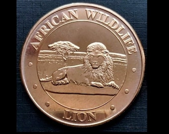 Peut inclure: Une pièce de monnaie de couleur bronze avec les mots "AFRICAN WILDLIFE" et "LION" en relief sur le bord. La pièce présente un relief détaillé d'un lion se reposant devant un arbre dans un paysage de savane.