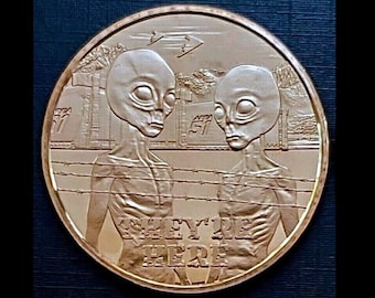 May include: A gold-coloured coin featuring a design of two grey alien figures standing behind a barbed wire fence. The text "They're Here" is inscribed on the coin.