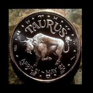 May include: A silver coin with a black background featuring the zodiac sign Taurus. The coin has the words "Taurus" and "April 21 - May 21" inscribed around the edge.