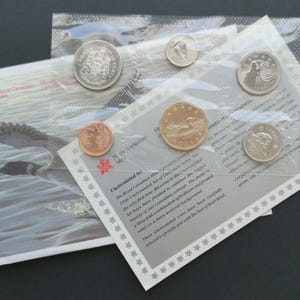 May include: A collection of Canadian coins in sealed plastic. The set includes various denominations, including a penny and a dollar coin. The packaging features text and a red maple leaf emblem.