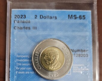 May include: A 2023 Canadian $2 coin featuring King Charles III in a gold-toned portrait on the obverse and a stylized maple leaf on the reverse. The coin is graded MS-65 and has a serial number of 128203.