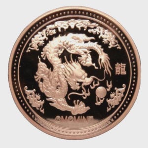 May include: A gold-coloured coin with a detailed relief of a dragon. The dragon is the central design, with the Chinese character for "dragon" visible. The coin has a dark background and the text "GMCMINT".