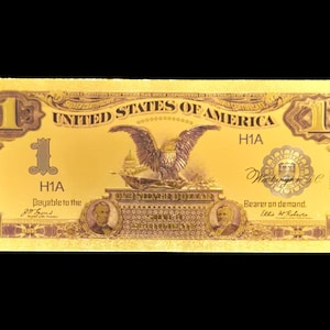 May include: A gold-coloured one-dollar bill with detailed design. The bill displays the words "United States of America" and "One Silver Dollar." An eagle with outstretched wings is centered above the words "One Silver Dollar."