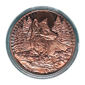 May include: A copper-colored coin featuring a detailed relief of a wolf's head, set against a backdrop of pine trees and a full moon. The coin is encased in a clear, protective casing with a blue-gray rim. The design evokes a wilderness theme.