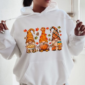 May include: White hoodie with a graphic of three gnomes wearing fall-themed outfits. The gnomes are holding a pie, a pumpkin, and a cup of coffee. The hoodie has a kangaroo pocket.