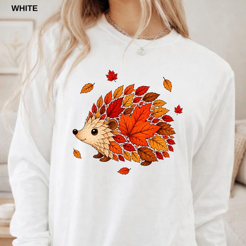 May include: A white long-sleeve shirt featuring a graphic of a hedgehog composed of various autumn-colored leaves in shades of red, orange, and brown. The hedgehog is shown in profile, with individual leaves forming its body and quills.