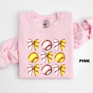 May include: Pink sweatshirt featuring a softball and bow design. The sweatshirt has a crew neck and long sleeves. The softball and bow design is repeated several times on the sweatshirt.
