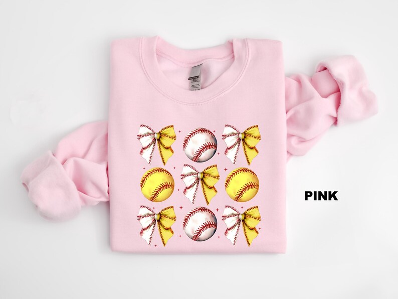 May include: Pink sweatshirt featuring a softball and bow design. The sweatshirt has a crew neck and long sleeves. The softball and bow design is repeated several times on the sweatshirt.