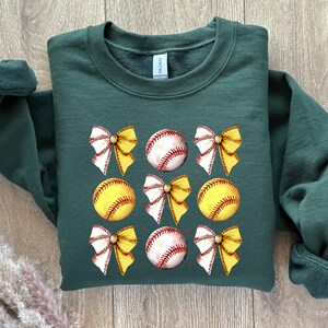 May include: A dark green sweatshirt featuring a design of yellow and white bows and baseballs. The baseballs are yellow with red stitching.