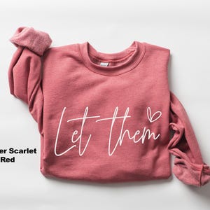 May include: Heather scarlet red sweatshirt with the words "Let them" in white script and a small heart. The long-sleeved top is folded, showing the inside of the cuffs. The sweatshirt is a solid color.