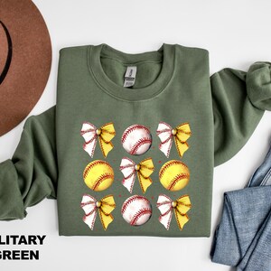 May include: A military green sweatshirt featuring a softball and bow design. The softball is yellow with red stitching and the bow is white with yellow accents.