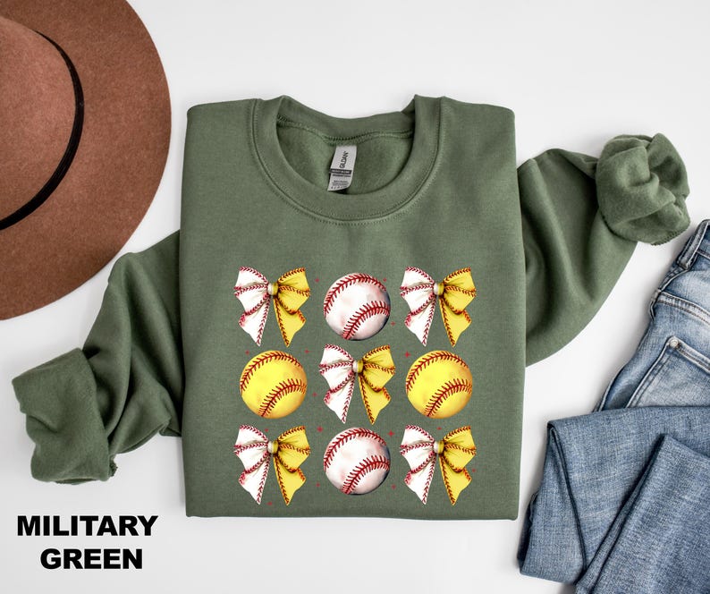 May include: A military green sweatshirt featuring a softball and bow design. The softball is yellow with red stitching and the bow is white with yellow accents.