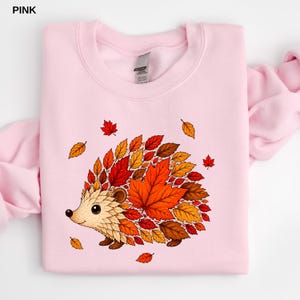 May include: A light pink crewneck sweatshirt featuring a graphic of a hedgehog whose body is composed of various autumn-colored leaves in shades of red, orange, and brown. The hedgehog has a cream-colored face and small dark eyes.