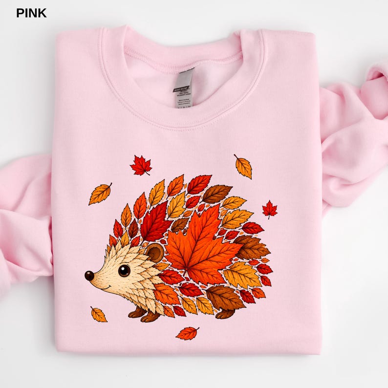 May include: A light pink crewneck sweatshirt featuring a graphic of a hedgehog whose body is composed of various autumn-colored leaves in shades of red, orange, and brown. The hedgehog has a cream-colored face and small dark eyes.