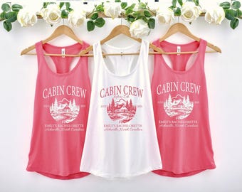May include: Three tank tops, two in coral and one in white, hang on wooden hangers. Each top features the text "CABIN CREW" above a graphic of a cabin and trees, with additional text below. A floral garland decorates the top.
