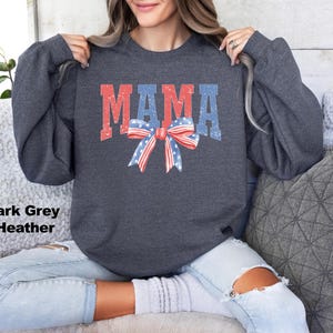 Pu&ograve; includere: Felpa color grigio scuro melange con la parola "MAMA" in lettere rosso, bianco e blu e un fiocco rosso, bianco e blu.