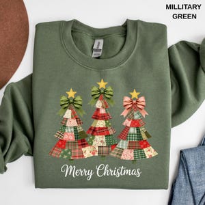 May include: A military green crewneck sweatshirt featuring a graphic of three Christmas trees made of patchwork fabric patterns. Each tree is topped with a gold star and a bow. The text Merry Christmas is printed in white below the trees.