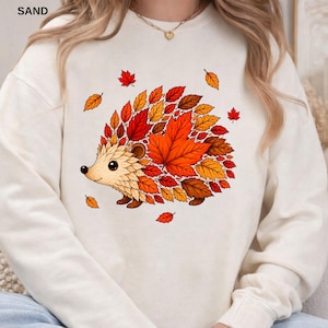 May include: A cream-colored crewneck sweatshirt featuring a graphic illustration of a hedgehog whose back is composed of vibrant autumn leaves in shades of orange, red, and brown. The design includes scattered falling leaves around the hedgehog.
