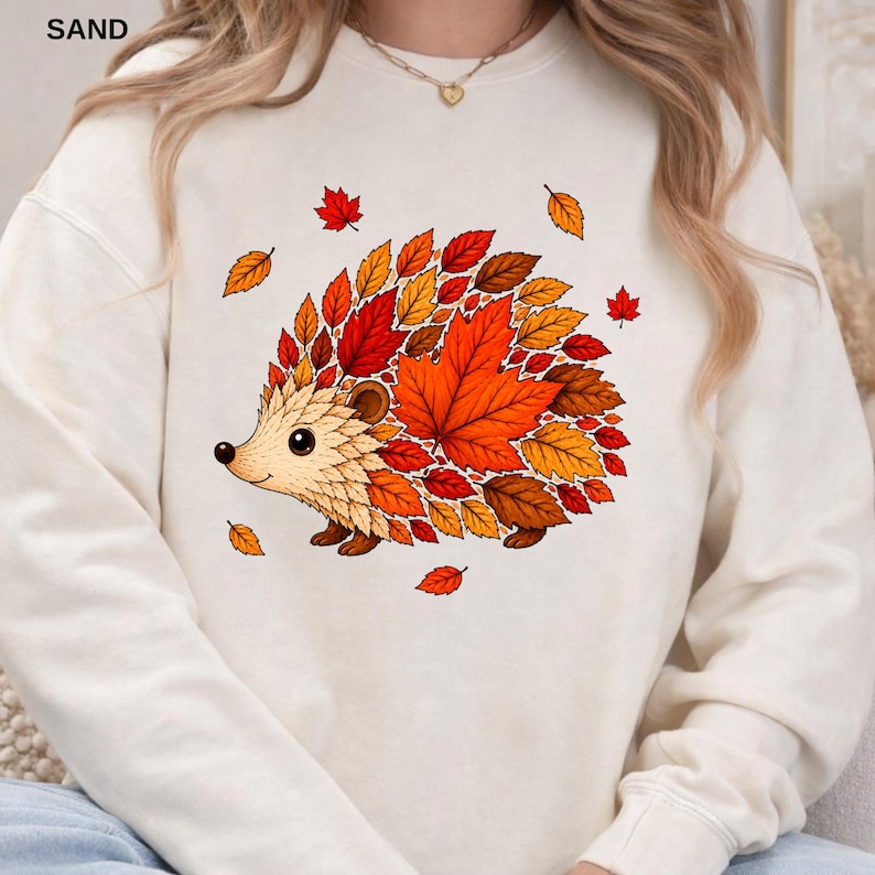 May include: A cream-colored crewneck sweatshirt featuring a graphic illustration of a hedgehog whose back is composed of vibrant autumn leaves in shades of orange, red, and brown. The design includes scattered falling leaves around the hedgehog.