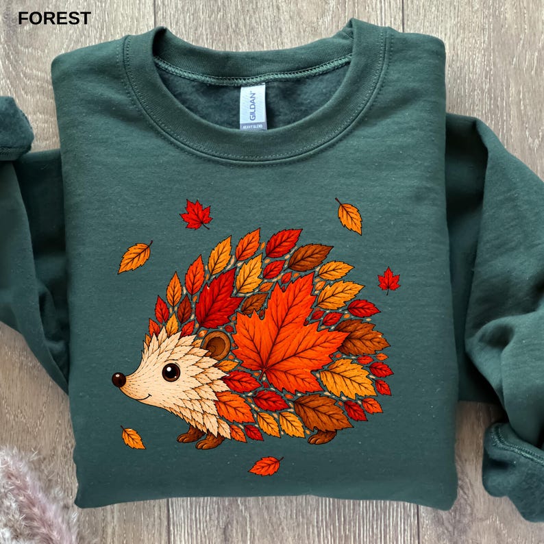 May include: A forest green crewneck sweatshirt featuring a graphic of a hedgehog whose body is composed of various autumn-colored leaves in shades of red, orange, and brown. The design includes scattered falling leaves around the hedgehog.