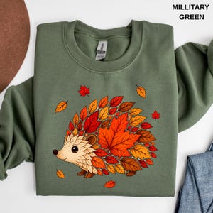Military green fall sweatshirt featuring a cute hedgehog surrounded by bright orange autumn leaves, pumpkins, and seasonal foliage, cozy woodland animal design for nature lovers and fall outfits.