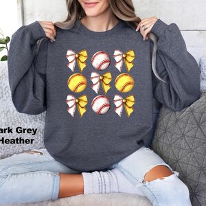 May include: Dark grey heather sweatshirt with a graphic of six yellow and white softball with yellow bows on a dark grey background.