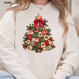 May include: A cream-coloured crewneck sweatshirt featuring a central graphic of a stylised Christmas tree composed of various patchwork fabric squares in red, green, and white patterns, topped with a gold star and a large red bow.