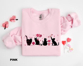 May include: Pink sweatshirt with a graphic of four black cats, each holding Valentine's Day themed items. The word "LOVE" is on the balloons. Heart-shaped sunglasses and lollipops are also in the image.