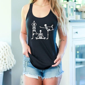 May include: Black tank top featuring a white graphic of three skeletons in yoga poses. The skeletons are in various positions, including standing, seated, and balancing poses. The tank top is sleeveless and made of a soft material.