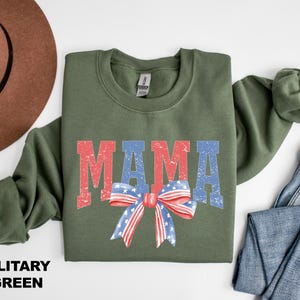 Pu&ograve; includere: Una felpa verde militare con la parola "MAMA" in lettere rosse, bianche e blu. Un fiocco rosso, bianco e blu con stelle &egrave; centrato sotto la parola "MAMA".