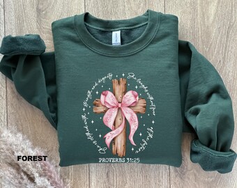 May include: A dark green sweatshirt featuring a white graphic of a wooden cross adorned with a pink ribbon. The text "She laughs without fear" and "clothed with strength and dignity" encircles the cross. The words "Proverbs 31:25" are printed beneath the cross.
