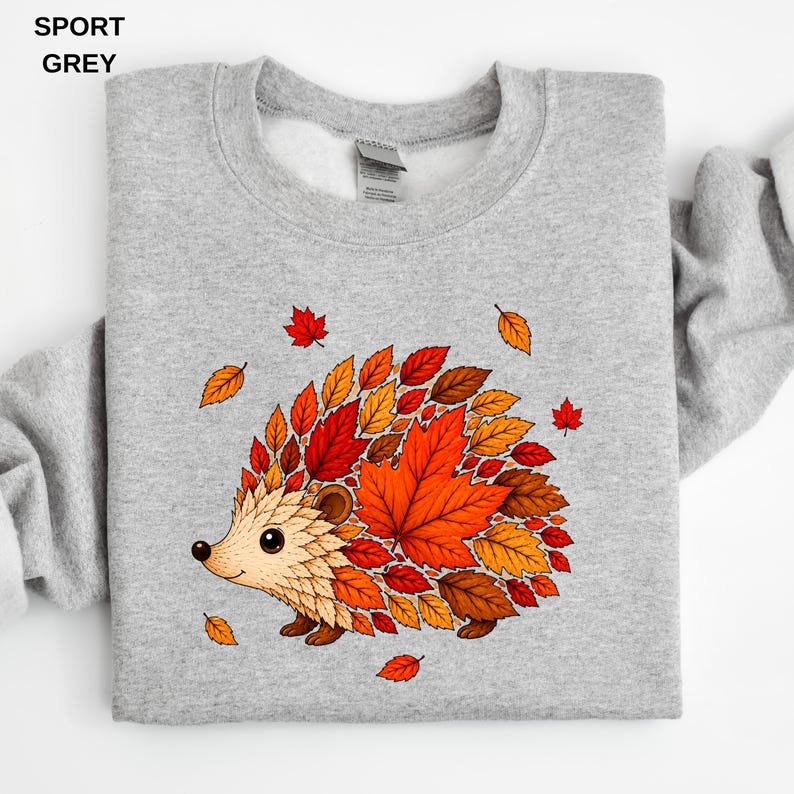 May include: A sport grey crewneck sweatshirt featuring a graphic of a hedgehog whose back is composed of various autumn-colored leaves in shades of red, orange, and brown. The text SPORT GREY is printed in the upper left corner of the image.