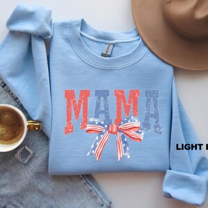 Pu&ograve; includere: Felpa blu chiaro con la parola "MAMA" in lettere rosse e blu con un fiocco rosso, bianco e blu con stelle al centro.