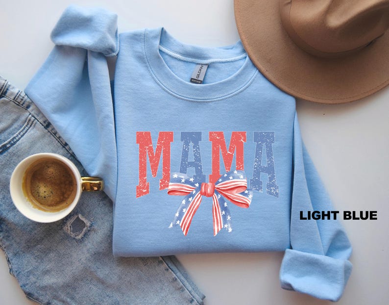 Pu&ograve; includere: Felpa blu chiaro con la parola "MAMA" in lettere rosse e blu con un fiocco rosso, bianco e blu con stelle al centro.