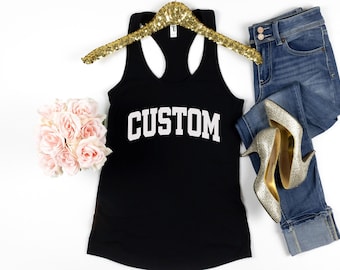 May include: Black tank top with the word "CUSTOM" in white block letters. The tank top is displayed with a gold sequined hanger, blue jeans, and silver high heels. A bouquet of pink roses is also in the image.