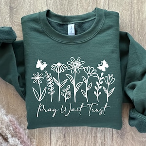 May include: A dark green sweatshirt with white floral and butterfly graphics. The words "Pray Wait Trust" are written in a script font below the flowers. The sweatshirt has a crew neck and long sleeves.