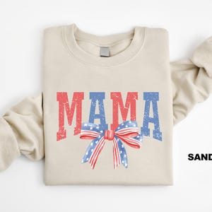 Pu&ograve; includere: Una felpa beige con la parola "MAMA" stampata in rosso, bianco e blu con un fiocco rosso, bianco e blu sotto. La felpa &egrave; piegata a met&agrave; e la parola "SAND" &egrave; stampata nell'angolo in basso a destra.