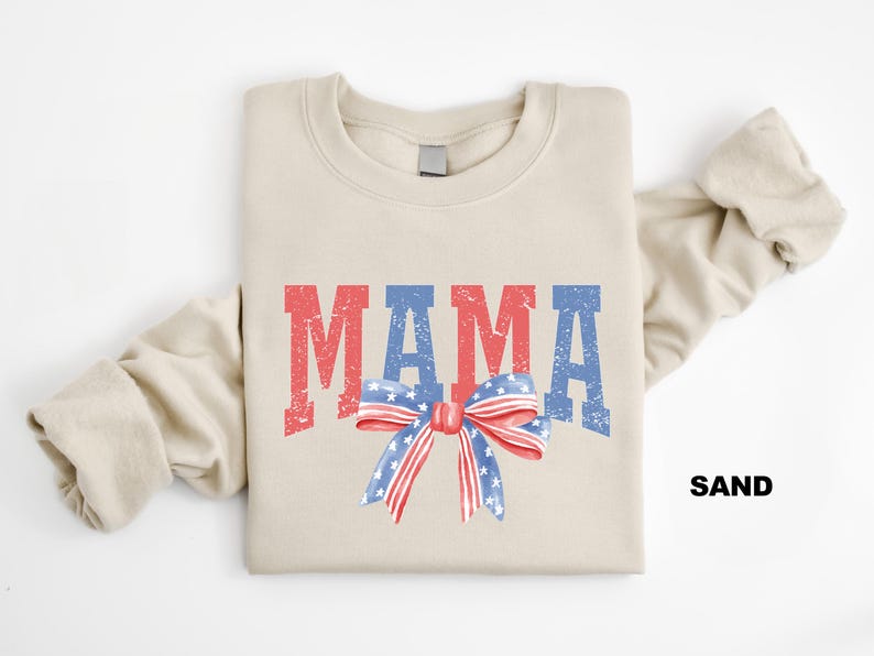 Pu&ograve; includere: Una felpa beige con la parola "MAMA" stampata in rosso, bianco e blu con un fiocco rosso, bianco e blu sotto. La felpa &egrave; piegata a met&agrave; e la parola "SAND" &egrave; stampata nell'angolo in basso a destra.
