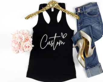 May include: A black racerback tank top with the word "Custom" in white script and a heart symbol. The tank top is displayed on a gold sequined hanger, next to a pair of blue jeans, and silver high heels. A bouquet of pink roses is also present.