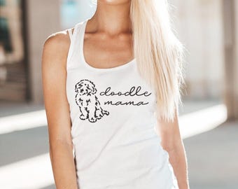 May include: White tank top with a black doodle dog graphic and the words "doodle mama". The model is wearing light wash denim jeans. The tank top has thin straps.