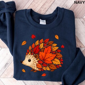 May include: A navy blue crewneck sweatshirt featuring a graphic of a hedgehog whose body is composed of various autumn leaves in shades of red, orange, yellow, and brown. The hedgehog has a light tan face and is surrounded by falling leaves.