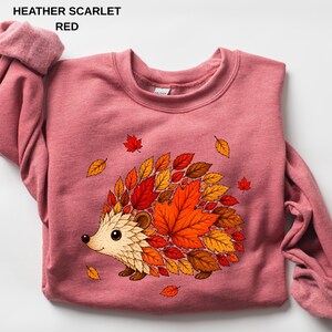 May include: A heather scarlet red crewneck sweatshirt featuring a graphic of a hedgehog whose back is composed of various autumn-colored leaves in shades of orange, yellow, and brown. The text HEATHER SCARLET RED is printed in the top left corner.