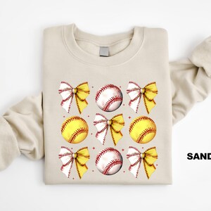 May include: A sand-coloured sweatshirt with a graphic design of yellow and white softball bows and baseballs. The text "SAND" is printed on the bottom right corner of the sweatshirt.