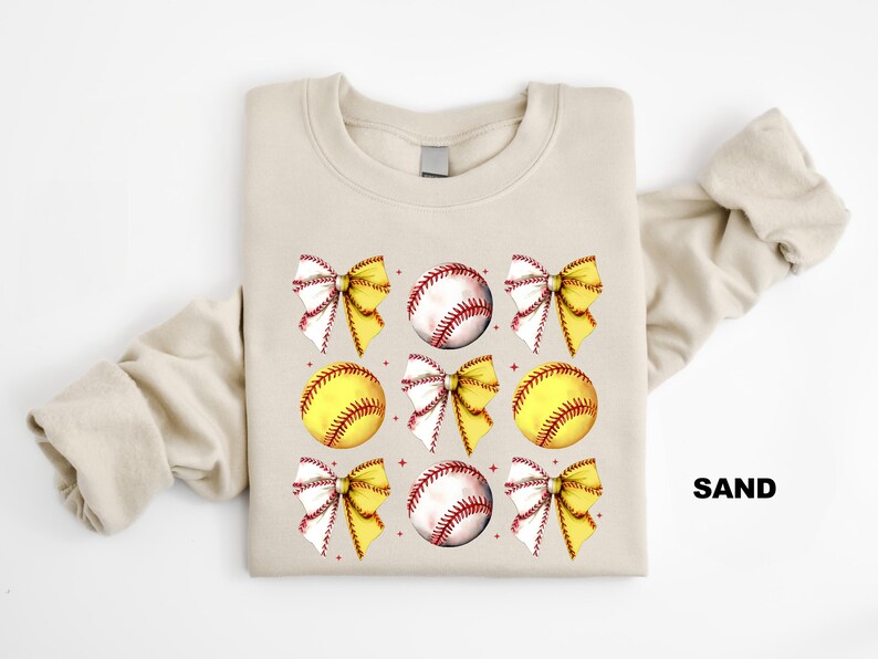 May include: A sand-coloured sweatshirt with a graphic design of yellow and white softball bows and baseballs. The text "SAND" is printed on the bottom right corner of the sweatshirt.