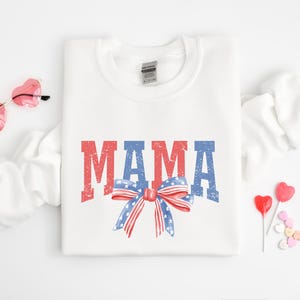 Pu&ograve; includere: Felpa bianca con la parola "MAMA" in lettere rosso, bianco e blu con un fiocco rosso, bianco e blu con stelle.
